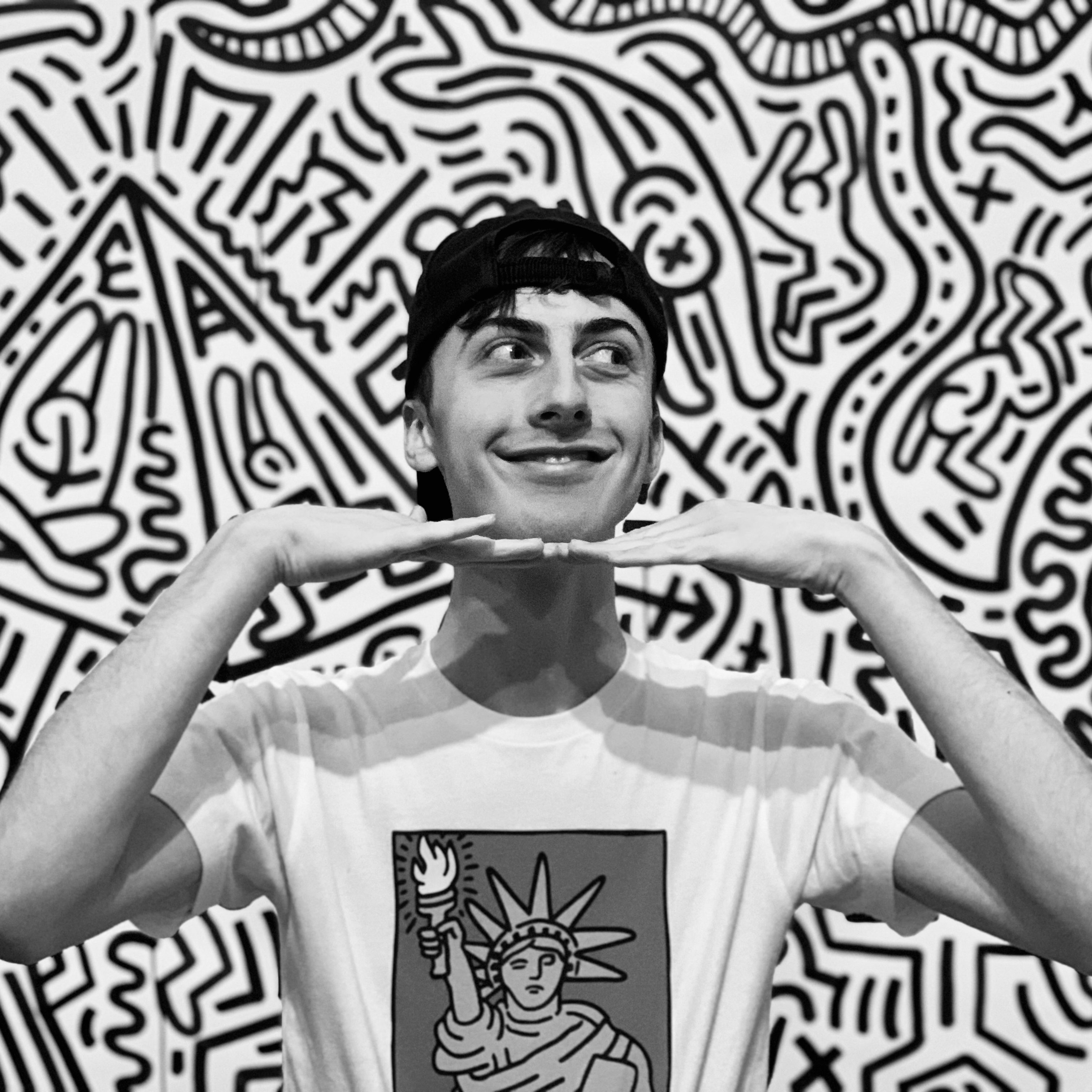 A black and white photo of Jamie standing in front of a Keith Haring mural. They are wearing a black baseball cap backwards and have a white t-shirt with another Keith Haring print on it 